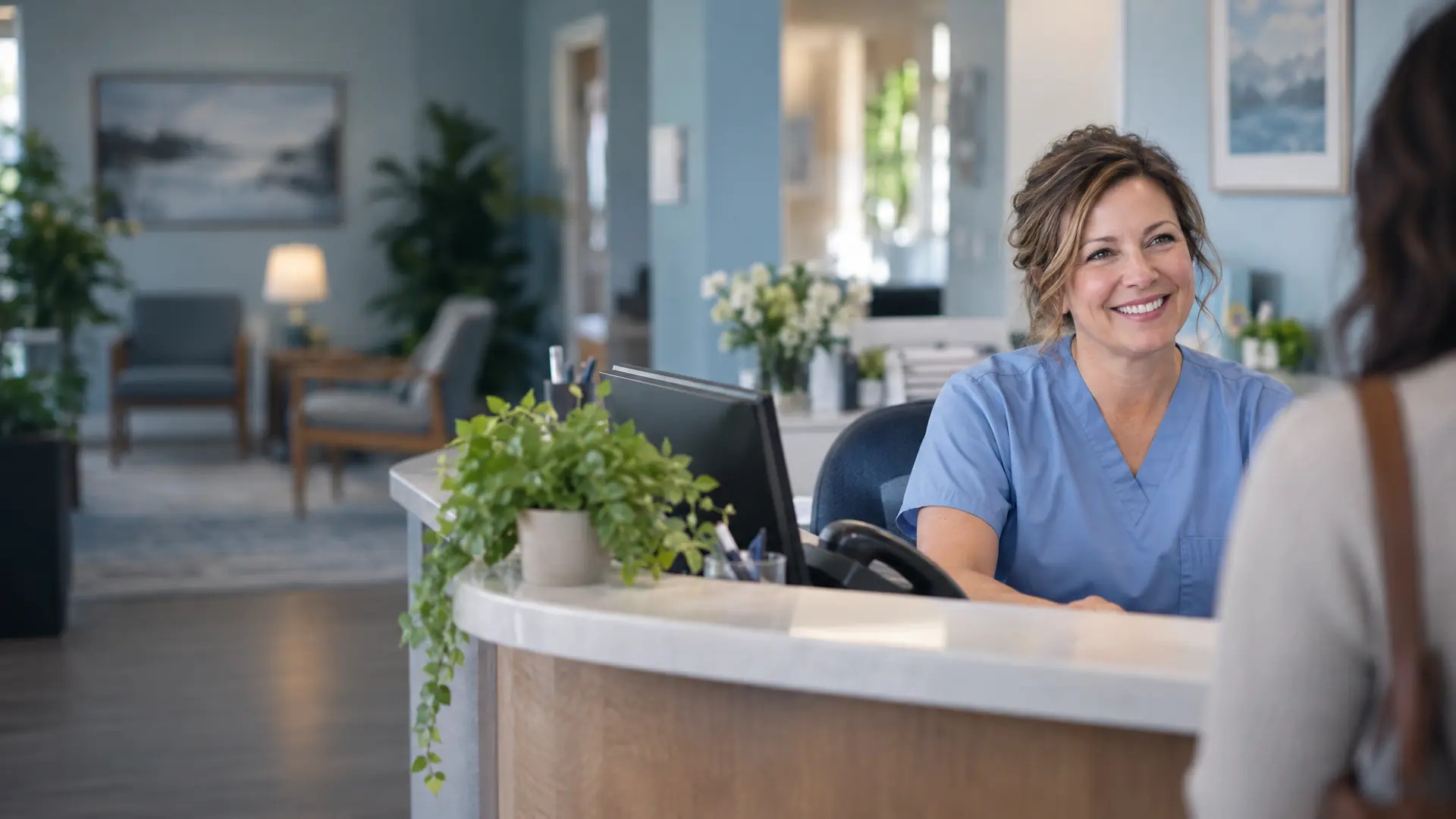 Medical front desk coordinator in blue scrubs smiling at an arriving patient at the reception desk of a bright family practice